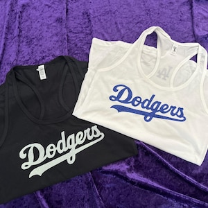 May include: Two tank tops, one black and one white, with the word "Dodgers" in script across the front. The black tank top has white lettering, and the white tank top has blue lettering. Both tops are on a purple background.