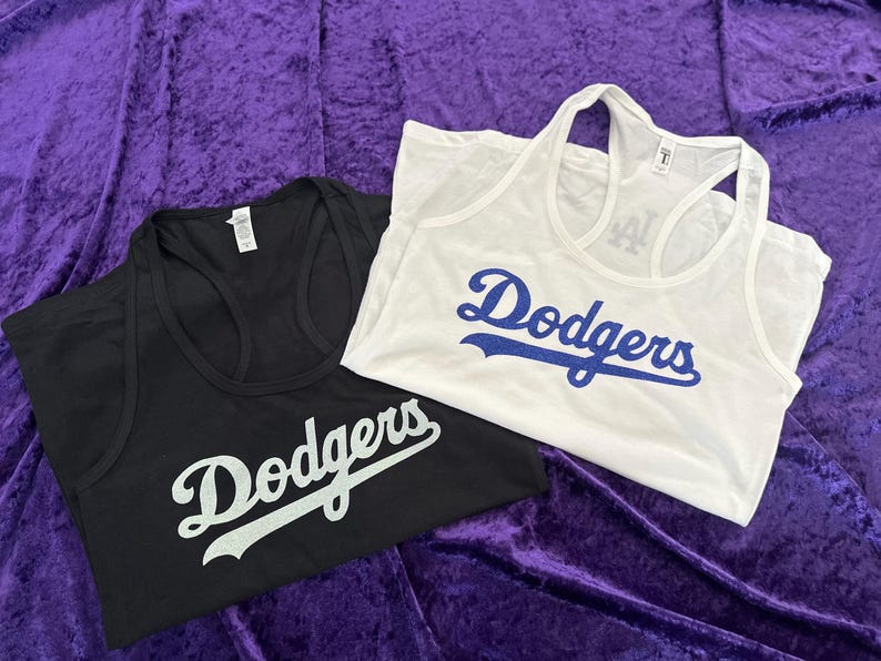 May include: Two tank tops, one black and one white, with the word "Dodgers" in script across the front. The black tank top has white lettering, and the white tank top has blue lettering. Both tops are on a purple background.