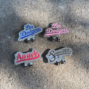May include: Four different golf hat clips with rhinestones. The clips feature the names "Los Dodgers", "Angels", "Serpientes", and "Los Dodgers" in different colours.