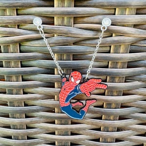 Puede incluir: Un collar con colgante de Spider-Man. El colgante presenta una figura de Spider-Man roja y azul con los brazos extendidos. El collar tiene una cadena plateada y está sujeto al colgante con pequeños bucles negros. El fondo es un patrón de mimbre tejido.
