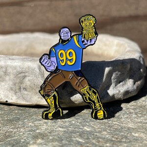 以下が含まれることがあります： マーベルコミックのキャラクター、サノスが、青と黄色のフットボールジャージを着て、背番号99番を付けています。彼は金のインフィニティ・ガントレットを掲げています。エナメル製のピンです。