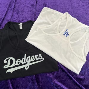 May include: Two tank tops, one black and one white, are displayed on a purple surface. The black tank top has the word "Dodgers" in white script. The white tank top has a blue "LA" logo.