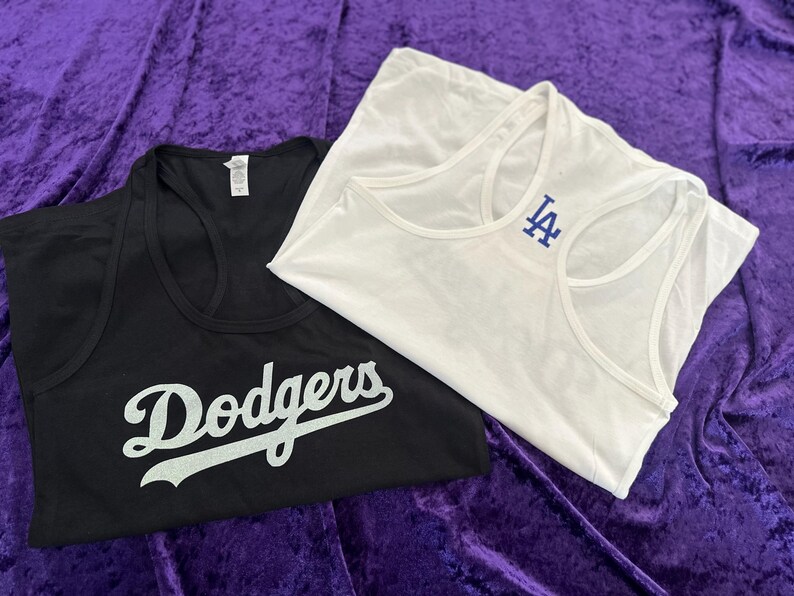 May include: Two tank tops, one black and one white, are displayed on a purple surface. The black tank top has the word "Dodgers" in white script. The white tank top has a blue "LA" logo.