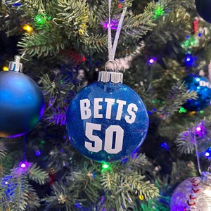 Puede incluir: Una bola de Navidad azul brillante con el nombre "BETTS" y el número "50" en letras blancas. El adorno cuelga de una cinta plateada en un árbol de Navidad con luces de colores y otros adornos.