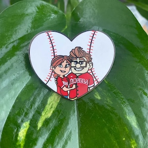 May include: A heart-shaped baseball pin with a cartoon couple inside. The man is wearing a red baseball jersey with "Los Angeles" written on it. The woman is wearing a red shirt with a baseball logo.