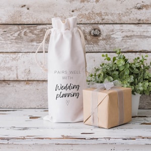 May include: White cotton wine bag with a black handwritten message that says "Pairs well with Wedding planning" and a small heart. The bag is sitting on a white wooden surface with a brown gift box and a potted green plant.