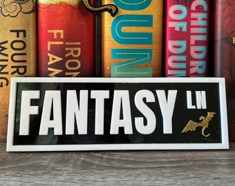 May include: A black and white sign with the word "FANTASY" in white letters and the letters "LN" in smaller white letters. A gold glitter dragon is on the right side of the sign. The sign is sitting on a wooden surface in front of a bookshelf with several fantasy novels.