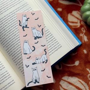 May include: A pink and white bookmark with a repeating pattern of white ghosts and black cats with bats flying around them. The bookmark has a watercolor effect.