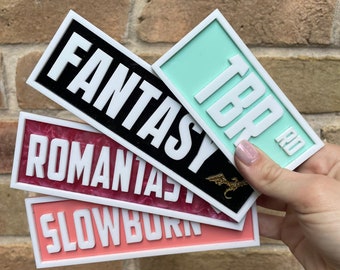 May include: Four rectangular signs with white lettering on different colored backgrounds. The top sign is turquoise with the word "TBR" in white. The second sign is black with the word "FANTASY" in white. The third sign is pink with the word "ROMANTASY" in white. The bottom sign is coral with the word "SLOWBURN" in white.