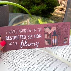 May include: A burgundy bookmark with white text that reads "I would rather be in the restricted section of the library". The bookmark features a hand reaching for a lantern in a bookshelf filled with books.