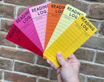 May include: A hand holds a fan of colorful reading logs against a brick wall. Each log is a different color: red, pink, orange, yellow, and light yellow. The words "READING LOG" are printed at the top of each log, with space for "Book Title" and "Rating".