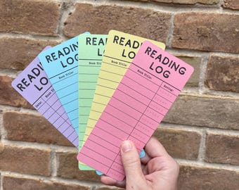May include: A hand holds a fanned stack of colorful reading log bookmarks. Each bookmark is a different pastel color: purple, blue, green, yellow, and pink. The bookmarks have the words "READING LOG" printed at the top, with spaces for book title, date, and rating.