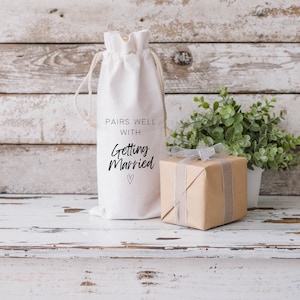 May include: White fabric wine bag with the text "Pairs well with getting married" and a small heart. A brown gift box with a white ribbon is next to the bag.