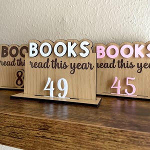 May include: Wooden book counter displays with the word "BOOKS" in white and pink, and the phrase "read this year" in brown script. Numbers 8, 49, and 45 are displayed in white and pink. The counters sit on a wooden shelf.