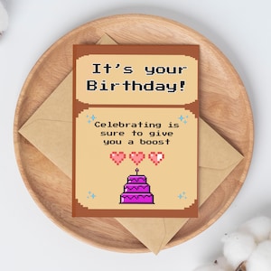 May include: A birthday greeting card with pixelated text that reads "It's your Birthday!" and "Celebrating is sure to give you a boost." The card features a pixelated cake and hearts. The card is on a wooden plate with an envelope.