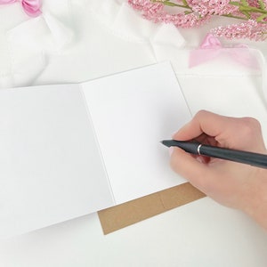 May include: A blank white greeting card is open, with a hand holding a black pen poised to write. The card rests on a brown paper base. Pink flowers and a pink bow are in the background, all on a white surface.