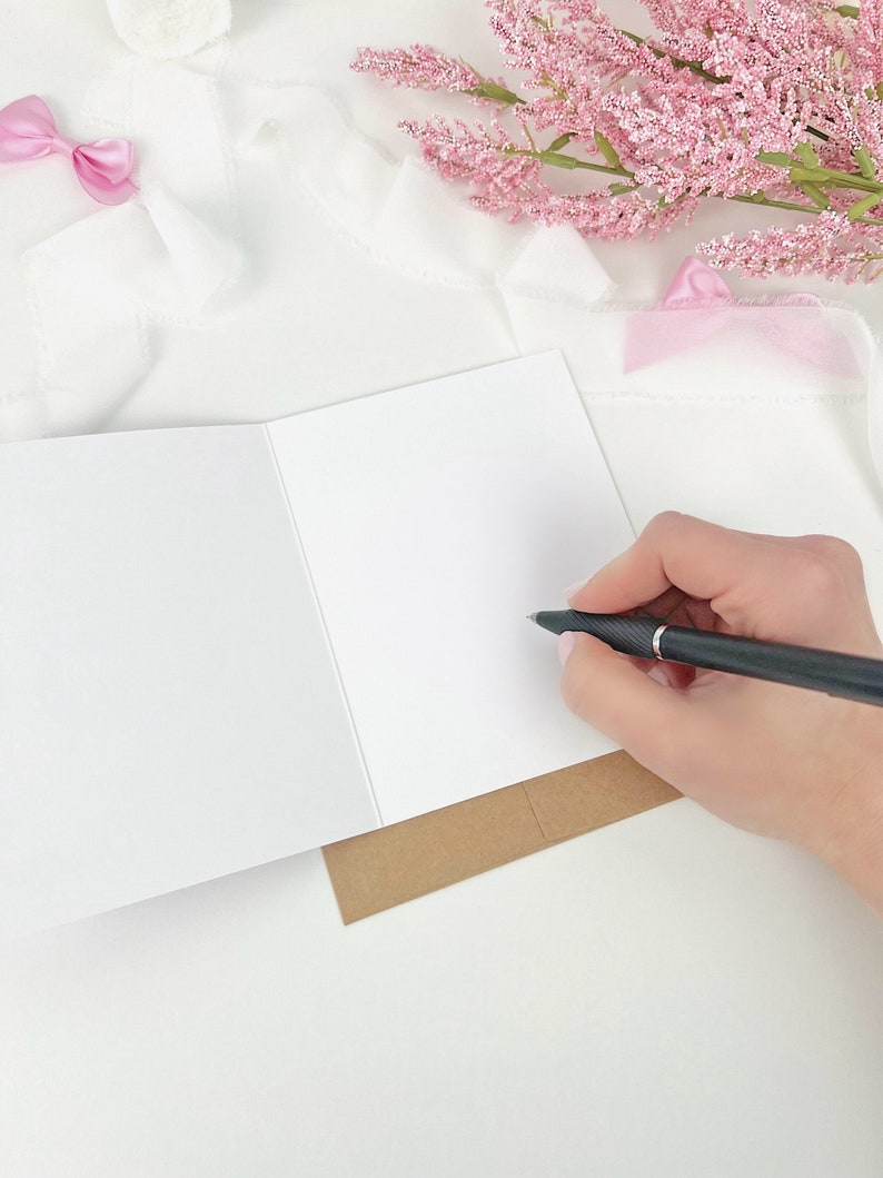 May include: A blank white greeting card is open, with a hand holding a black pen poised to write. The card rests on a brown paper base. Pink flowers and a pink bow are in the background, all on a white surface.