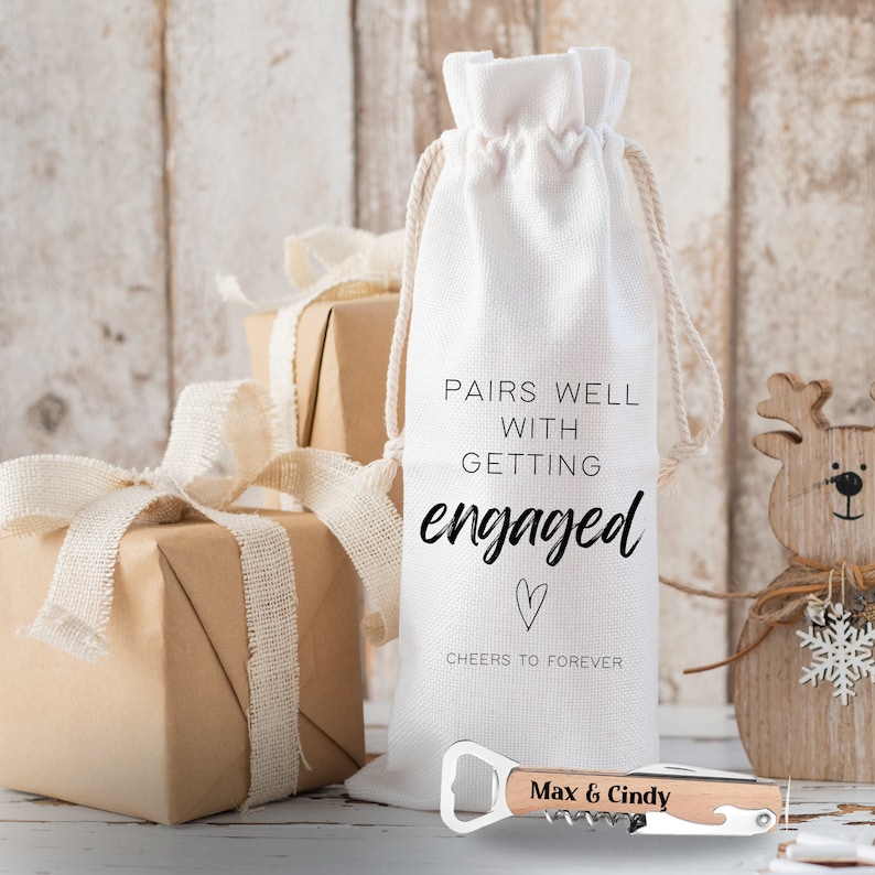 May include: White fabric wine bag with the text "Pairs well with getting engaged" and a heart. A wooden bottle opener with the text "Max & Cindy" is in the foreground.