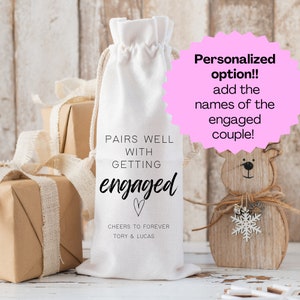 May include: White fabric wine bag with the text "Pairs well with getting engaged" and a heart. The bag also has the text "Cheers to forever Tory & Lucas".