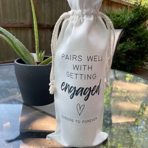 May include: White fabric wine bag with black text that reads "Pairs well with getting engaged" and a heart symbol. The text below the heart says "Cheers to forever".