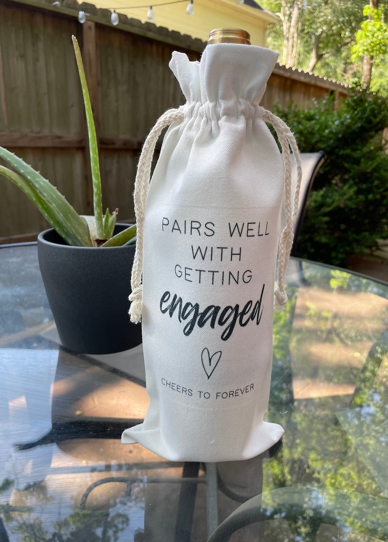 May include: White fabric wine bag with black text that reads "Pairs well with getting engaged" and a heart symbol. The text below the heart says "Cheers to forever".