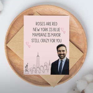 May include: A greeting card with the text "Roses are red, New York is blue, Mamdani is mayor, still crazy for you." The card features a photo of a man in a suit and a skyline of New York City. The card is on a wooden plate with an envelope.