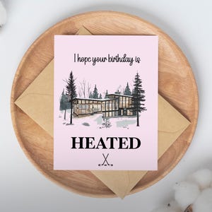 May include: A pink birthday card with the text "I hope your birthday is HEATED" and a graphic of a house in a snowy landscape. The card is on a wooden tray with a brown envelope.