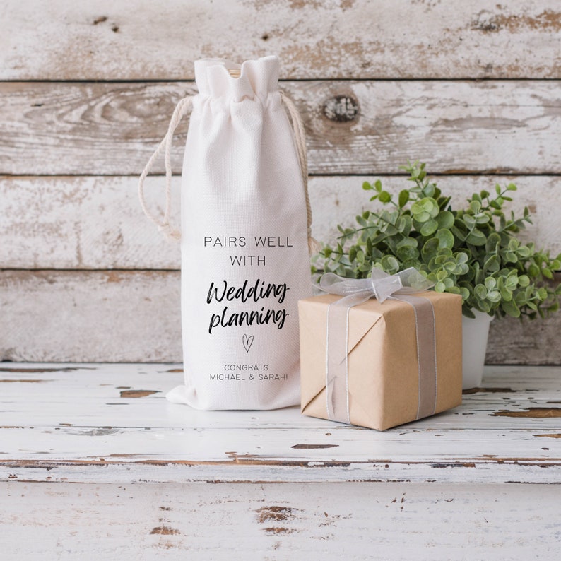 May include: White fabric wine bottle bag with the text "Pairs well with Wedding planning" and a heart symbol. The text "Congrats Michael & Sarah!" is below the heart symbol. A small brown gift box with a white ribbon is in the foreground.