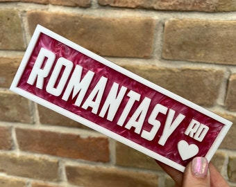 May include: A white sign with the words "ROMANTASY RD" in white lettering with a white heart symbol. The sign is on a pink and white marbled background.
