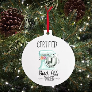 May include: White round Christmas ornament with a red ribbon. The ornament features a blue and silver stand mixer with floral accents and the text "CERTIFIED Bad Ass BAKER".