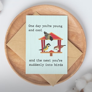 May include: A greeting card with the text "One day you're young and cool and the next you're suddenly into birds." The card features a birdhouse with three illustrated birds. The card is on a wooden tray with a brown envelope.