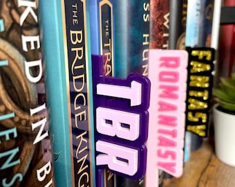 May include: A collection of books with colorful spines, featuring decorative bookmarks. The bookmarks are labeled "TBR", "ROMANTASY", and "FANTASY" in bold, contrasting colors. The books are on a wooden shelf.