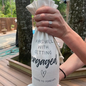 May include: White fabric wine bottle bag with the text "First Well With Getting Engaged" and a heart symbol. The text "Cheers to Forever" is below the heart symbol.
