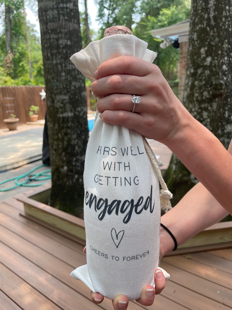 May include: White fabric wine bottle bag with the text "First Well With Getting Engaged" and a heart symbol. The text "Cheers to Forever" is below the heart symbol.
