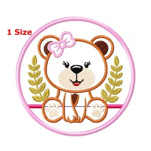Pode incluir: Desenho bordado de um urso marrom com um laço rosa sentado em um círculo com folhas verdes. O desenho está sobre um fundo branco com um círculo rosa ao redor do urso.