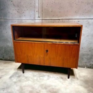 May include: A mid-century modern wooden cabinet with a two-door lower section and an open shelf above. The cabinet has a warm, reddish-brown finish and dark legs. The doors have a small, dark handle.