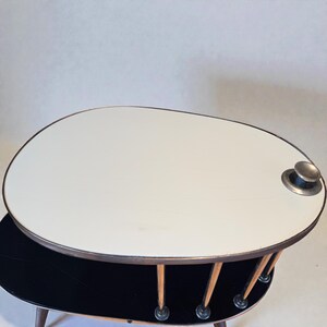 May include: A mid-century modern coffee table with an oval, two-tiered design. The top tier is white with a metal rim and a silver knob. The lower tier is black. The table has wooden legs and a unique, retro aesthetic.