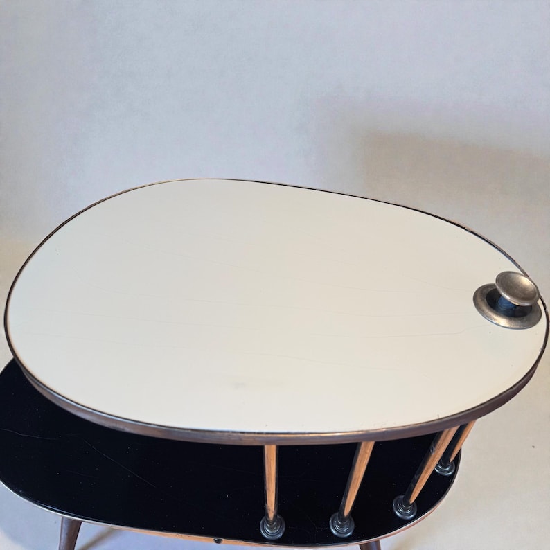 May include: A mid-century modern coffee table with an oval, two-tiered design. The top tier is white with a metal rim and a silver knob. The lower tier is black. The table has wooden legs and a unique, retro aesthetic.