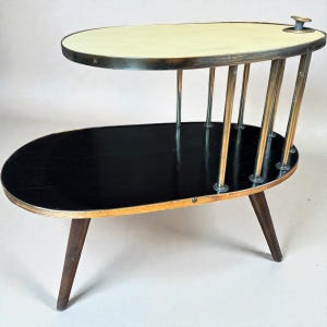 May include: A mid-century modern side table with an oval shape. The top tier is cream-colored, and the lower tier is black. The table has brass-colored metal supports and dark wooden legs. The table is approximately 24 inches long.