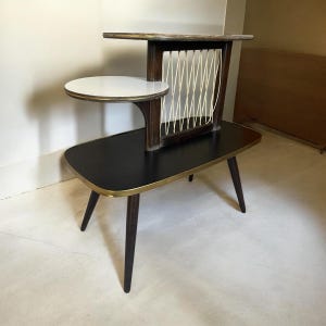 Könnte beinhalten: Ein moderner Beistelltisch aus der Mitte des Jahrhunderts mit einer schwarzen Tischplatte und goldenen Verzierungen. Der Tisch hat ein einzigartiges Design mit einem weißen Regal und einer geflochtenen Drahtgitterplatte.