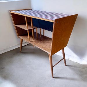 May include: A mid-century modern wooden cabinet with a striped top. The cabinet has angled sides, shelves, and slender legs. The top surface features red and white stripes. The cabinet is made of light brown wood and has a dark blue interior.