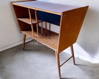 May include: A mid-century modern wooden cabinet with a striped top. The cabinet has angled sides, shelves, and slender legs. The top surface features red and white stripes. The cabinet is made of light brown wood and has a dark blue interior.