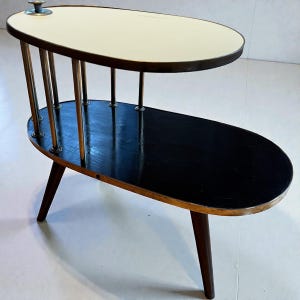 May include: A mid-century modern two-tiered side table. The top tier is oval with a white surface, while the lower tier is black. The table has brass-colored supports and dark wooden legs. The table is approximately 28 inches long.