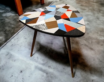 May include: A mid-century modern side table with a geometric pattern in shades of gray, blue, red, and brown. The table has a triangular top and three legs.