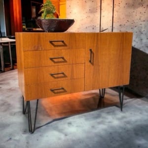 May include: A wooden cabinet with four drawers and a door, featuring black metal hairpin legs. The cabinet has a warm, natural wood finish and rectangular drawer pulls. A decorative bowl with greenery sits on top.