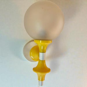 May include: A vintage wall sconce with a frosted globe shade. The fixture features a bright yellow, hourglass-shaped body with a white accent band. The sconce is mounted on a white and yellow circular base, creating a retro aesthetic.