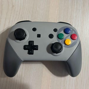 May include: A gray and black Nintendo Switch controller with a retro design. The controller has a gray face with black buttons and a black d-pad. The buttons are labeled A, B, X, Y, and Start. The controller has a gray grip on each side.