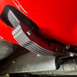 May include: A black and gray automotive component, possibly a protective shield or trim piece, is mounted against a bright red surface. The component has a layered design with gray and black stripes. Fasteners secure it to the vehicle's underside.
