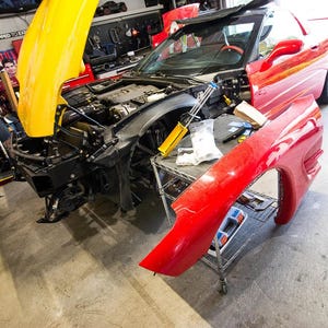 May include: A red sports car with its hood and front fender removed, revealing the engine and internal components. The hood is yellow. The car is in a garage setting, suggesting repair or customization.