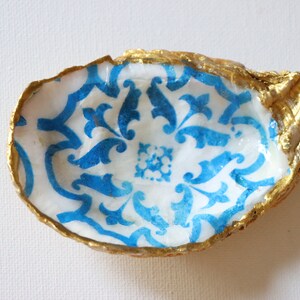 May include: A small, white oyster shell dish with a blue and white patterned design. The shell is painted gold around the edges.